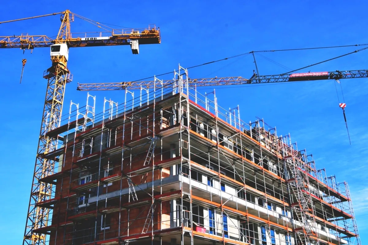 Building under construction wrapped in scaffolding, with two tower cranes against a blue sky